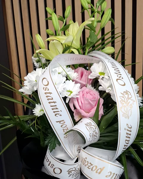 Wiązanka pogrzebowa na liściach: Wiązanka pogrzebowa z białymi liliami, różowymi różami i białymi chryzantemami, ułożona na dużych liściach i trawie ozdobnej, przewiązana szarfą z napisem Ostatnie pożegnanie. Kwiaciarnia Bukieteria Gorzów Wielkopolski