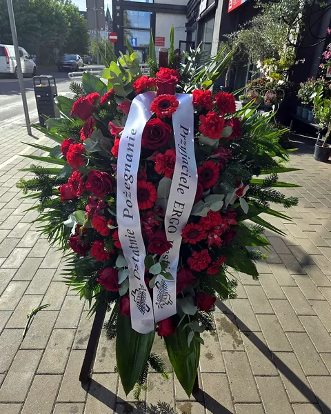 Wiązanka pogrzebowa na świerku z czerwonych kwiatów: Duża wiązanka na podkładzie ze świerku, ułożona z czerwonych róż, goździków, gerber i alstromerii, uzupełniona eukaliptusem i liśćmi palmy, z białą szarfą. Kwiaciarnia Bukieteria Gorzów Wielkopolski